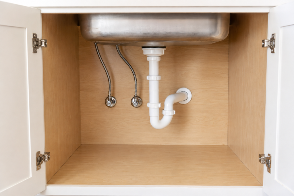 Empty sink cabinet showing supply valves, hoses, drain joints, and the cabinet floor.